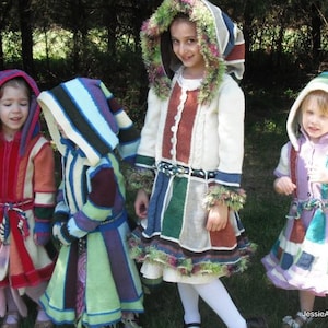 May include: Four children wearing colorful, patchwork-style hooded coats and dresses. The garments feature a variety of colors including red, orange, green, blue, and purple. The children are standing outside in front of a green background.