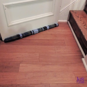 Puede incluir: Un tope de puerta artesanal con un patrón de cuadros azul marino, azul y beige. El tope cilíndrico descansa contra una puerta blanca sobre un suelo de madera. El tope mide aproximadamente 91 cm de largo.