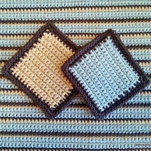以下が含まれることがあります： ベージュと水色の、濃い茶色の縁取りが付いた正方形のかぎ針編みコースター2つ。縞模様のブランケットの上に置かれています。コースターは手作りで、テクスチャードステッチパターンを示しています。ブランケットは、青、ベージュ、茶色の水平ストライプが特徴です。