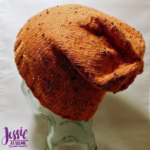 以下が含まれることがあります： 手編みのオレンジ色のビーニー帽。まだら模様のデザイン。柔らかく、質感のある糸で作られた帽子で、ゆったりとしたフィット感です。透明なマネキンの頭に展示されています。