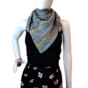 Può includere: Uno scialle lavorato a maglia grigio e multicolore di forma triangolare. Lo scialle presenta un motivo a maglia strutturato con dettagli traforati lungo un bordo. La tavolozza dei colori comprende sfumature di grigio, giallo e rosa.