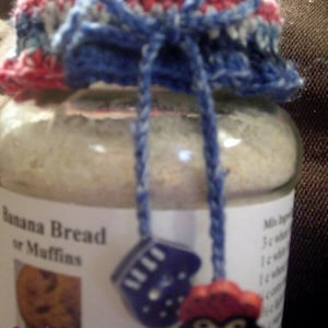 May include: A glass jar with a white label that reads "Banana Bread or Muffins". The jar is topped with a blue and red crocheted lid with a blue ribbon tied around it. The jar also has a blue and white crocheted mitten and a red and white crocheted penguin attached to it.
