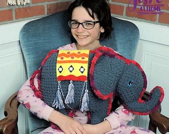 Op de afbeelding: Een handgemaakt grijs olifantenkussen met rode en gele accenten. De olifant heeft een decoratieve deken met kwastjes en een blauw oog. Het kussen wordt vastgehouden door een persoon met een bril en een roze pyjama.