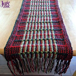 May include: A knitted table runner with a red, green, white, and black checkered pattern. The runner has a fringe on the bottom edge.