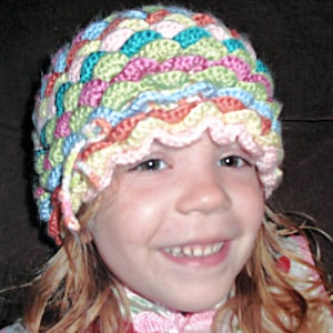 May include: A young person wearing a colourful crocheted hat with a scalloped edge. The hat is made with a variety of pastel colours, including pink, blue, green, and yellow.