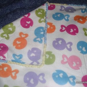 May include: A white fleece blanket featuring a repeating pattern of colorful fish with heart-shaped tails. The fish are in shades of pink, orange, green, blue, and purple. The blanket has a crocheted edge in pastel colors.