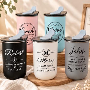 May include: Five cylindrical stainless steel tumblers with black lids and light blue flip-top closures. The tumblers come in black, white, grey, pink, and light blue, each featuring custom printed names, job titles, and decorative graphic elements.
