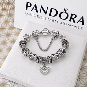 Op de afbeelding: Een zilveren bedelarmband met diverse textuurkralen, hartvormige bedels en kleine rode steentjes, voorzien van een centrale hartvormige hanger en een veiligheidsketting, geplaatst voor een witte doos met de tekst PANDORA.