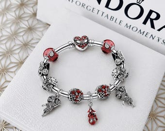 Pode incluir: Uma pulseira de prata com vários berloques temáticos do Homem-Aranha em esmalte vermelho e preto com pedras transparentes. Os berloques incluem uma máscara em forma de coração, contas redondas estampadas e figuras pendentes numa caixa.