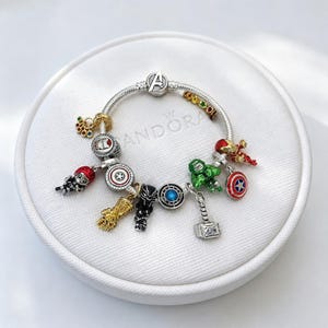 Op de afbeelding: Een zilveren bedelarmband met diverse Marvel-bedels, waaronder een gouden infinity gauntlet, een groen Hulk-figuurtje, een zilveren hamer van Thor, een zwart pantermasker, een rood-blauw schild en een sluiting met het Avengers-logo aan een zilveren ketting.