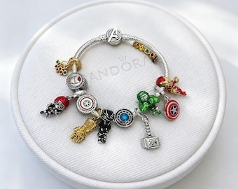 Pode incluir: Uma pulseira de prata com vários berloques temáticos da Marvel, incluindo uma manopla do infinito dourada, uma figura verde do Hulk, um martelo do Thor prateado, uma máscara de pantera negra, um escudo vermelho e azul e um fecho com logo Avengers.