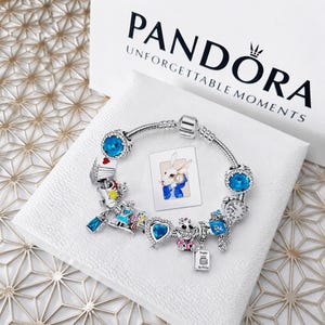 Può includere: Un bracciale in argento con vari charm a tema Alice nel Paese delle Meraviglie, tra cui un cuore blu, un orologio, una carta da gioco e lo Stregatto. Il bracciale è esposto su una scatola bianca con il testo PANDORA UNFORGETTABLE MOMENTS.