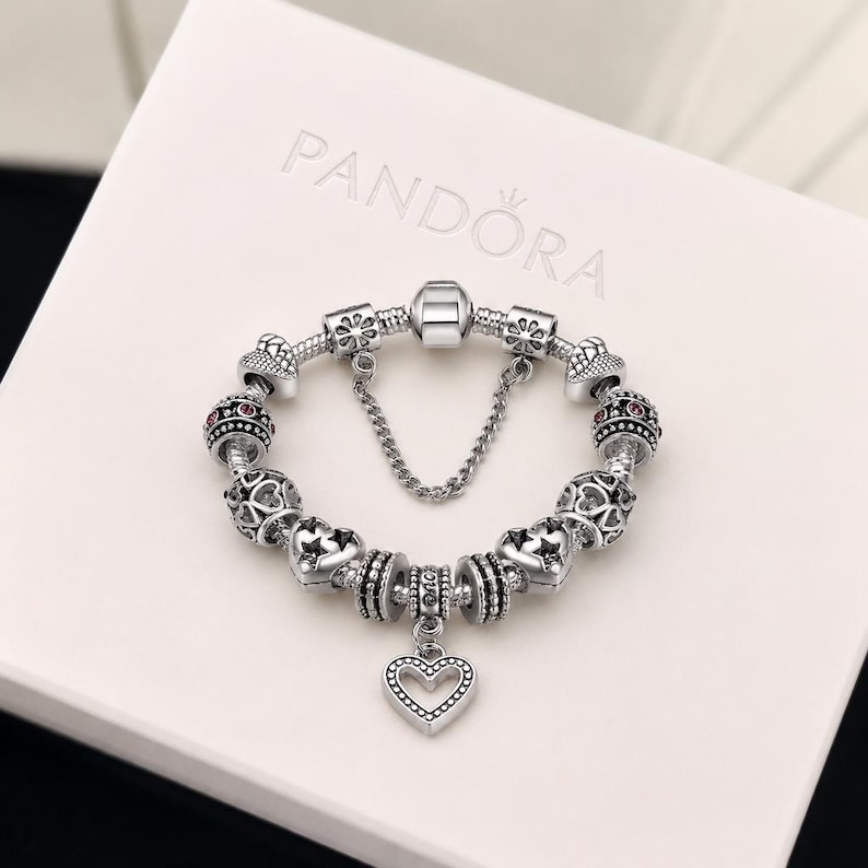 Pode incluir: Uma pulseira de prata com v&aacute;rios berloques texturizados, pingentes em forma de cora&ccedil;&atilde;o e uma corrente de seguran&ccedil;a, sobre uma caixa branca com o logotipo da Pandora em relevo na tampa. A pulseira inclui separadores e contas com padr&otilde;es.