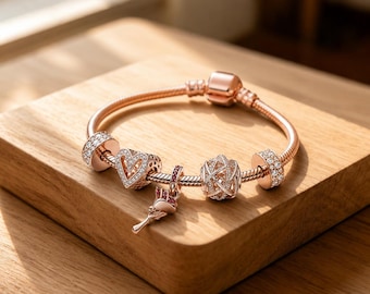 Pode incluir: Uma pulseira de berloques em ouro rosa com várias contas decorativas, incluindo um berloque em forma de coração com pedras transparentes, um berloque pendente de rosa com detalhes em rosa, uma conta esférica vazada e dois espaçadores cilíndricos.