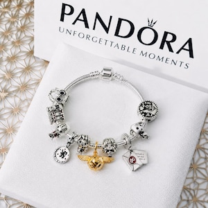 May include: A silver Pandora charm bracelet featuring various Harry Potter-themed charms, including a gold-toned snitch, a letter, and character figures. The bracelet is displayed in front of a white Pandora box with the brand name and logo printed in black.