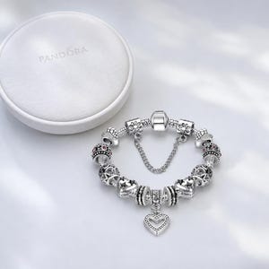 Pode incluir: Uma pulseira de berloques em prata com v&aacute;rias contas intrincadas, incluindo designs em forma de cora&ccedil;&atilde;o e pequenas pedras cor-de-rosa, com um pendente de cora&ccedil;&atilde;o central e uma corrente de seguran&ccedil;a. Uma caixa de joias redonda branca Pandora.