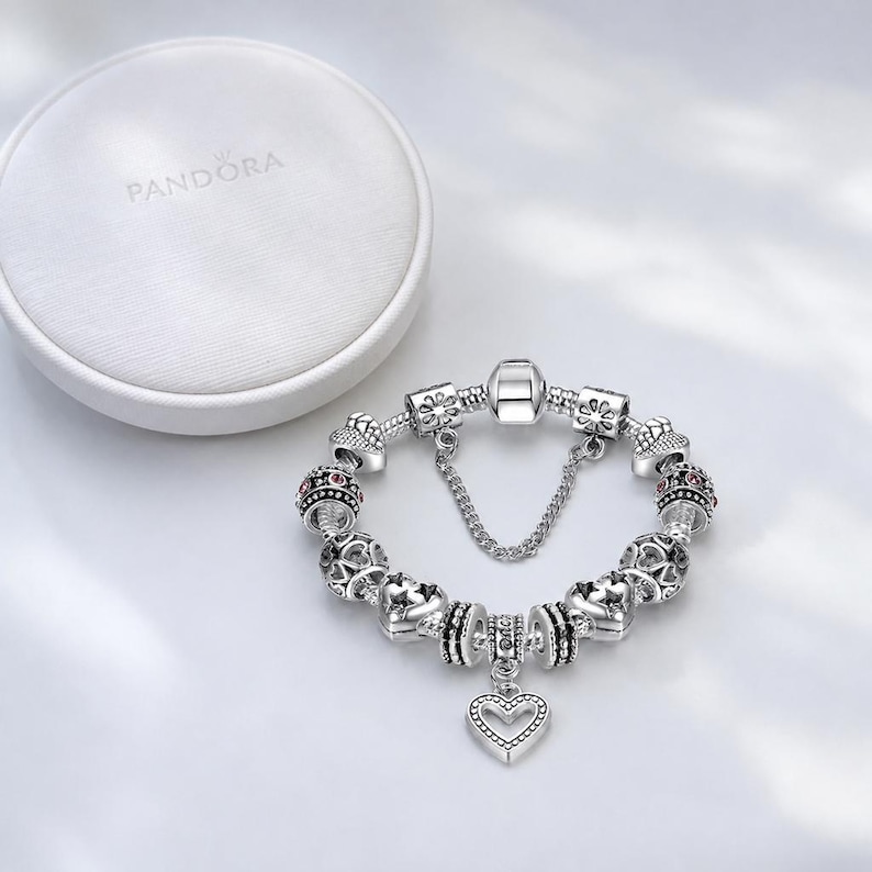 Pode incluir: Uma pulseira de berloques em prata com v&aacute;rias contas intrincadas, incluindo designs em forma de cora&ccedil;&atilde;o e pequenas pedras cor-de-rosa, com um pendente de cora&ccedil;&atilde;o central e uma corrente de seguran&ccedil;a. Uma caixa de joias redonda branca Pandora.