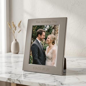 May include: A silver-toned rectangular picture frame featuring the engraved text Our Wedding Day at the top and 09.05.2026 at the bottom. The frame holds a photograph of a couple in wedding attire, with the groom in a suit and the bride in a lace dress.