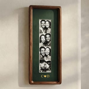 Puede incluir: Un marco de fotos rectangular vertical de madera con un paspartú verde oscuro. El marco contiene una tira de cuatro fotografías en blanco y negro que muestran a una pareja en varias poses. Debajo de las fotos, las letras doradas C y D flanquean un corazón.