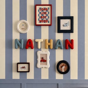 May include: A wall display featuring the name NATHAN in three-dimensional, multi-coloured letters. Surrounding the name are six framed pictures, including illustrations of a goose, a toy car, a plane, and a sign reading LONG LIVE BOYHOOD.