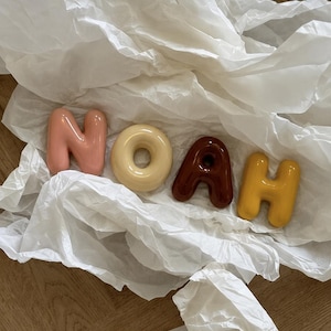 May include: Four individual, glossy, three-dimensional letters spelling the name NOAH. The letter N is light pink, the O is cream, the A is dark brown, and the H is mustard yellow. Each letter has a smooth, rounded, balloon-like appearance.