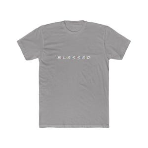 May include: A grey short-sleeved t-shirt featuring the word BLESSED printed in white capital letters across the chest. Between each letter, there are small, colourful dots in red, blue, and yellow, mimicking a classic television show logo style.
