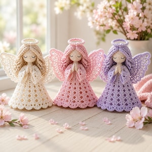 May include: Three crocheted angel figurines standing side by side. Each angel features a round head with closed eyes, a halo, textured hair, and large wings. The angels are dressed in long, flared dresses with a diamond-patterned crochet design.