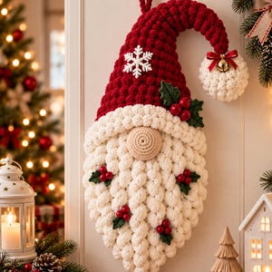 Peut inclure: Une décoration murale en crochet représentant un gnome avec un grand bonnet rouge orné d'un flocon blanc, un nez beige rond et une longue barbe blanche. Le bout du bonnet porte un pompon blanc avec une clochette dorée et un nœud rouge.