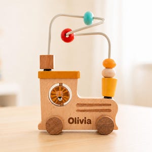May include: A wooden train-shaped bead maze toy featuring a lion illustration in a circular window, the name Olivia printed on the side, and a wire frame with teal, red, yellow, and natural wooden beads for sliding and tactile play.