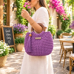 May include: A purple crocheted handbag with a round wooden handle and a matching purple tassel. The bag features a small rectangular wooden tag with the word Handmade engraved on the front, attached to the textured, thick-stitched fabric.