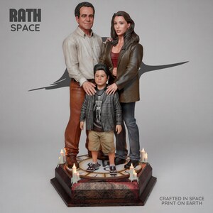 May include: A three-person family figurine set standing on a circular base with clock markings and small candles. The adult figures wear a beige shirt with brown pants and a brown blazer with dark pants. The child wears a gray jacket and tan shorts.