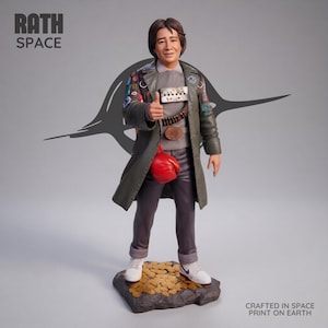 May include: A figurine of a person with dark hair wearing a grey shirt, dark trousers, and a long olive green coat adorned with various patches. The figure gives a thumbs up, wears a red boxing glove, and stands on a base of gold coins.
