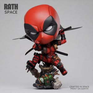 Könnte beinhalten: Eine stilisierte Deadpool-Figur in dynamischer Action-Pose mit rot-schwarzem Anzug, zwei Katanas und einer kleinen Handfeuerwaffe. Der Sockel enthält ein grünes Fass mit der Aufschrift BOOM. Der Text RATH SPACE und CRAFTED IN SPACE PRINT ON EARTH ist zu sehen.