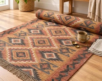 May include: A rectangular area rug featuring a repeating geometric diamond pattern in shades of rust orange, mustard yellow, and charcoal gray. The rug has a fringed edge and is partially rolled, revealing a ceramic mug and folded cloth.