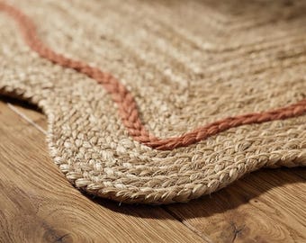 May include: A woven natural fiber rug featuring a scalloped edge and a prominent, curved terracotta-colored braided border detail. The rug is constructed from thick, textured tan fibers arranged in a dense, concentric pattern.