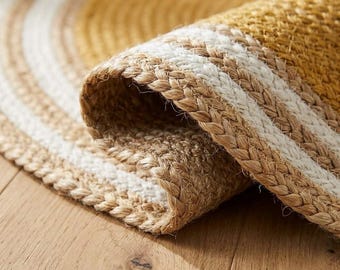 May include: A round braided rug featuring concentric circles of natural tan jute fibers interspersed with bands of white. The edge of the rug is folded over, revealing the thick, textured weave of the braided material and the circular pattern.