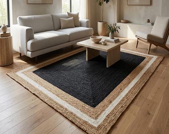 May include: A rectangular area rug featuring a large black center panel, a wide white border, and an outer border made of natural tan woven jute fibers. The rug is positioned on a light-colored wooden floor in a living room setting.