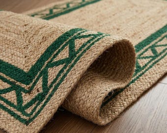 May include: A rectangular woven rug made of natural tan fibers featuring a dark green border with a repeating geometric triangular pattern. The rug is partially rolled, revealing the textured, braided construction of the material.