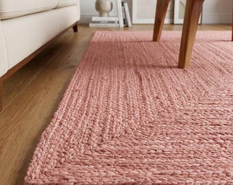 May include: A rectangular area rug featuring a dusty rose pink color and a thick, braided texture. The rug is constructed from woven natural fibers, displaying a consistent, chunky knit pattern that covers the entire surface of the floor covering.