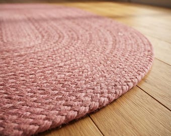 May include: A dusty rose oval rug featuring a thick, braided texture. The rug is constructed from tightly woven fibers that create a consistent, repeating pattern across the entire surface, showcasing a soft, muted pink color throughout.