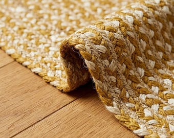 May include: A woven area rug featuring a braided texture with alternating strands of mustard yellow and off-white fibers. The rug is partially folded over, revealing the thick, interlaced construction of the natural material fibers throughout.