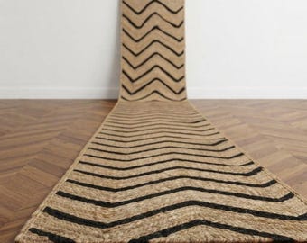 May include: A long, rectangular runner rug featuring a natural tan fiber texture with a repeating black chevron pattern. The rug extends from the foreground into the background, displaying a consistent zigzag design across its entire surface.