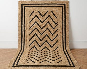 May include: A rectangular area rug crafted from natural tan woven fibers, featuring a bold black chevron pattern in the center and a thick black border frame. The rug is displayed standing upright against a wall, showcasing its textured surface.