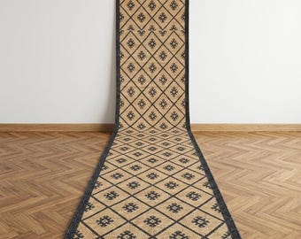 May include: A long, rectangular runner rug featuring a natural tan fiber base with a repeating black diamond grid pattern. Each diamond contains a small, centered black geometric motif. The rug has a thick, solid black border.