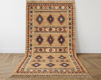 May include: A rectangular woven jute rug featuring a natural tan base with repeating geometric patterns in deep blue and rust red. The design includes diamond shapes and horizontal bands, finished with a fringe along the top and bottom edges.
