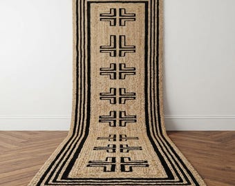 May include: A long rectangular runner rug made of natural tan woven fibers. The rug features a bold black border and a central vertical column of repeating black geometric symbols that resemble stylized T-shapes arranged in pairs.