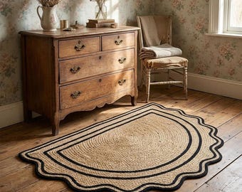 May include: A semi-circular jute rug featuring a natural tan woven texture with a thick black border and a scalloped edge. The rug is placed on a wooden floor in front of a wooden dresser and a chair, showcasing a distinct curved design.