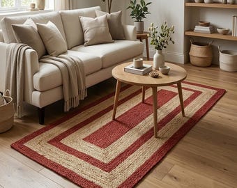 May include: A rectangular area rug featuring a woven texture with alternating concentric borders of natural tan and deep red. The rug is positioned on a light-colored wooden floor, extending from beneath a round wooden coffee table.