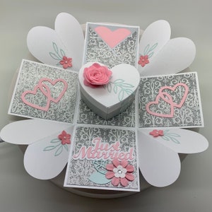 May include: A handmade wedding-themed explosion box. The box is white and silver with pink accents, including a heart-shaped box with a felt rose on top. The box is decorated with hearts, flowers, and the words "Just Married".