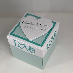 May include: A light blue gift box with a white lid and a heart-shaped topper. The lid features the word "LOVE" in teal. The heart-shaped topper has the names "Caroline & Colton" and the date "February 10, 2019".